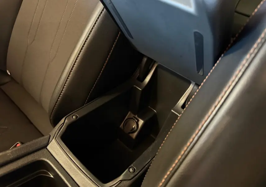 Gros plan sur la console centrale ouverte avec prise 12V, sièges noirs et surpiqûres marron visibles dans une CUPRA LEON ST blanche 2024.