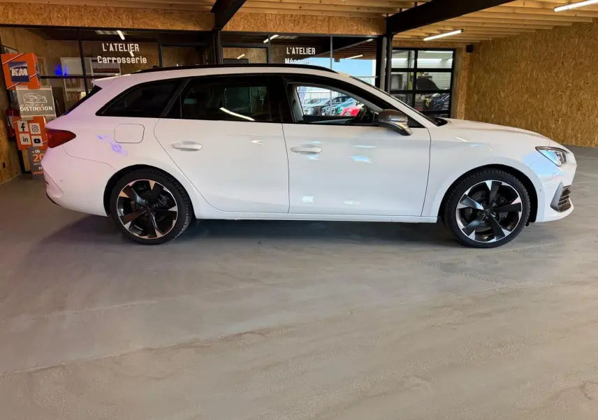 Vue de profil côté gauche d'une CUPRA Leon ST blanche 2024 avec jantes noires et toit vitré en intérieur d'atelier.