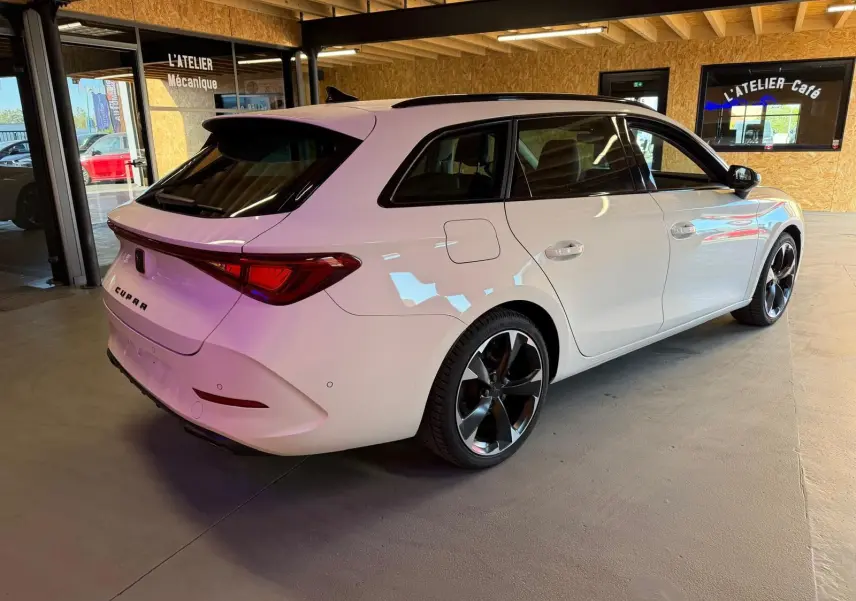 Vue 3/4 arrière droite d'une CUPRA Leon ST blanche 2024 avec jantes noires brillantes et toit panoramique en intérieur d'atelier.