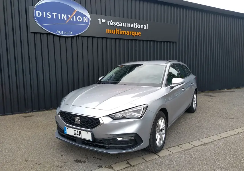 SEAT Leon ST gris en 3/4 avant droit, garée devant un mur noir avec un panneau Distinxion visible.