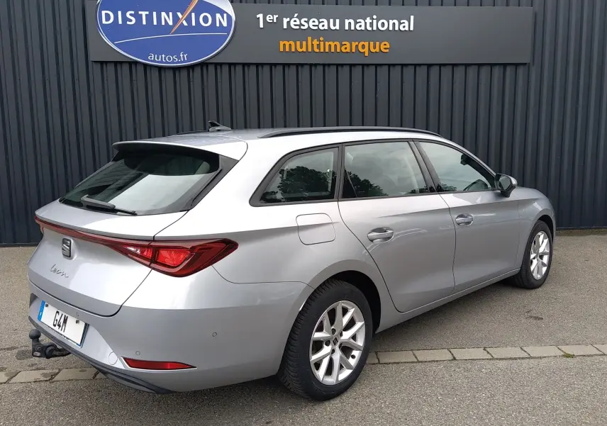 Vue 3/4 arrière droite d’une SEAT LEON ST gris métallisé avec attelage, garée devant un mur gris.