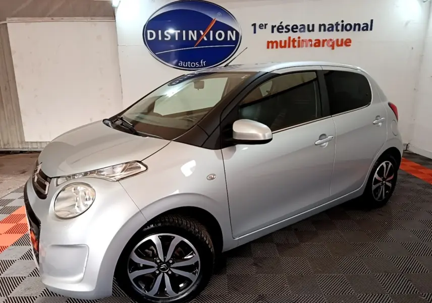 Citroën C1 gris clair vue 3/4 avant droit en intérieur, avec jantes noires et logo Distinxion en arrière-plan.
