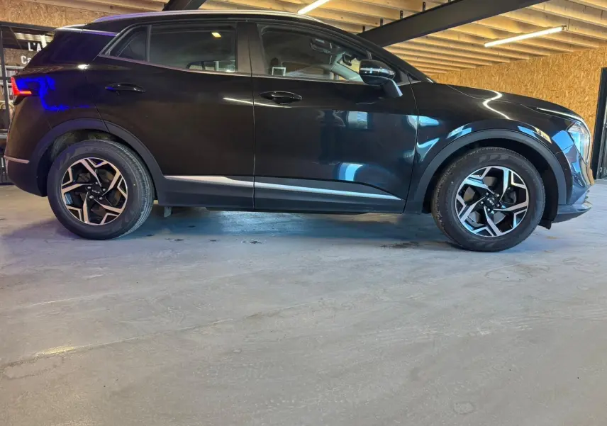 Profil côté gauche d’un KIA Sportage noir 2022 avec jantes alliage et détails chromés sous un plafond en bois.