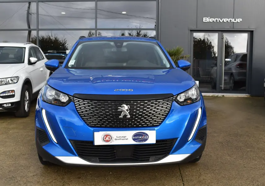 Vue frontale d'un Peugeot 2008 bleu 2021 avec calandre noire et phares LED, devant un garage avec enseigne "Bienvenue".