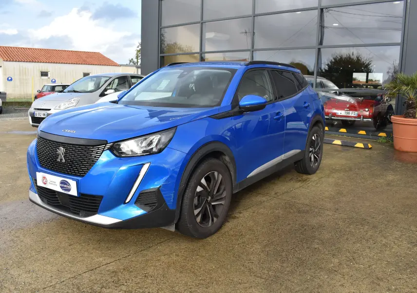 Peugeot 2008 bleu en 3/4 avant droit, avec calandre noire et jantes alliage sombres, stationné devant un bâtiment vitré.
