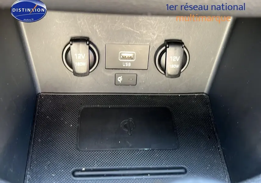 Gros plan sur la console centrale de la Hyundai i30 2024 montrant prises 12V, port USB et chargeur sans fil noir.