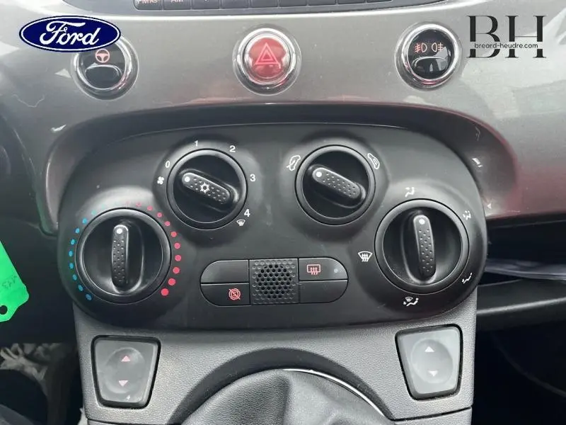 Gros plan sur la console centrale noire d’une Fiat 500 marron de 2010, montrant les commandes de climatisation et les boutons d’alerte.