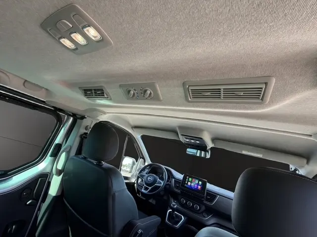 Vue intérieure avant du Nissan Primastar gris clair 2025, montrant le volant multifonctions, écran tactile central et toit avec aérations.