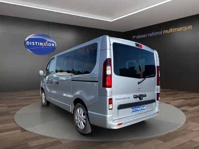 Vue 3/4 arrière droite d’un Nissan Primastar gris clair avec vitres teintées et porte latérale visible en showroom.