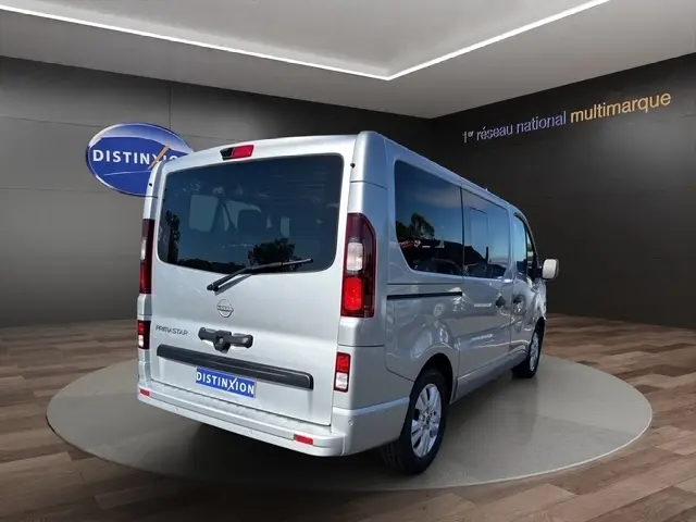 Vue 3/4 arrière droite d’un Nissan Primastar gris clair avec vitres teintées et porte latérale visible en showroom.
