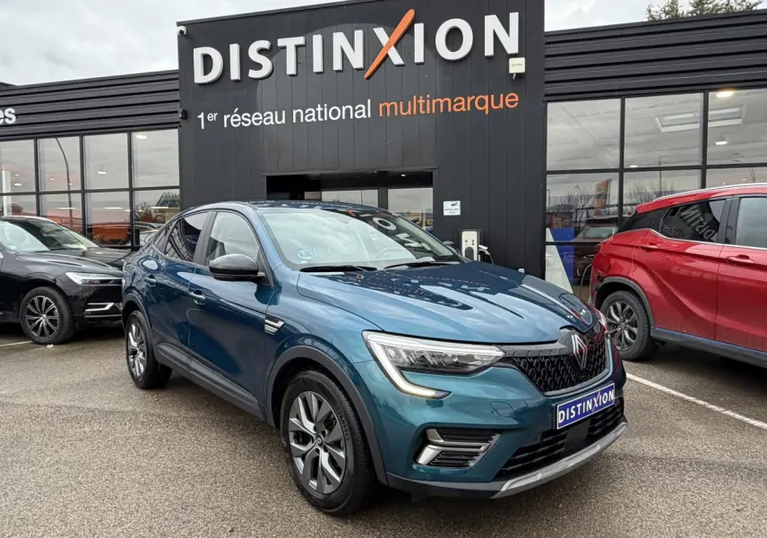 Vue 3/4 avant d'une Renault Arkana bleu Zanzibar métallisé avec jantes alliage grises, stationnée devant un showroom Distinxion.