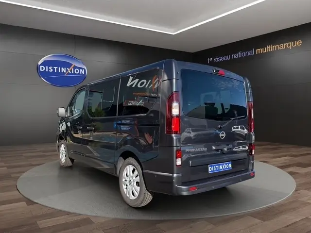 Vue 3/4 arrière droite d'un Nissan Primastar gris foncé avec vitres teintées et jantes argentées sur fond showroom.