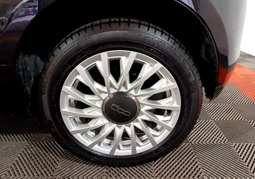Gros plan sur la roue avant droite d'une FIAT 500 noire, mettant en valeur la jante argentée au design étoilé.