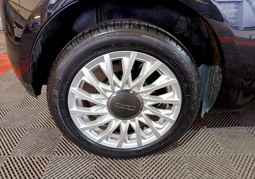 Gros plan sur la roue avant droite d'une FIAT 500 noire, mettant en valeur la jante argentée au design complexe.