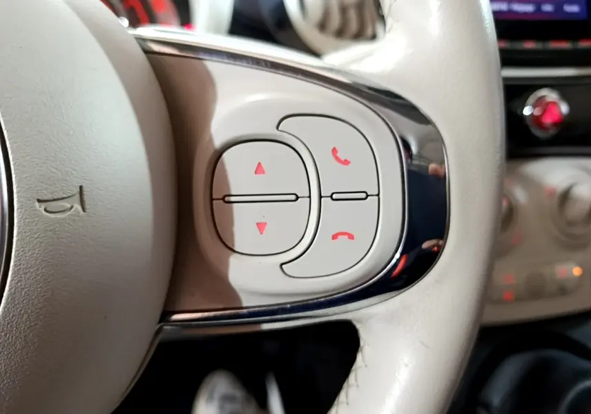 Gros plan sur les commandes blanches du volant cuir beige d'une Fiat 500 Lounge 2019, avec tableau de bord flou en arrière-plan.