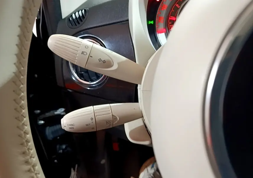 Gros plan sur les commandes de clignotants et régulateur au volant beige d’une Fiat 500 Lounge noire, tableau de bord partiellement visible.