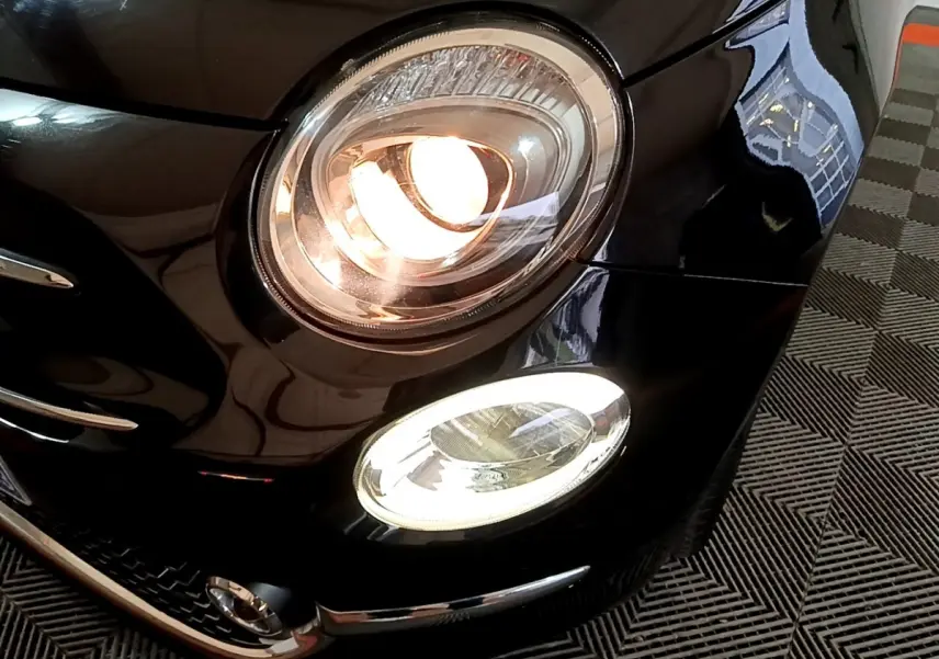 Gros plan sur le phare avant droit allumé d'une Fiat 500 noire, mettant en valeur ses feux LED et détails chromés.