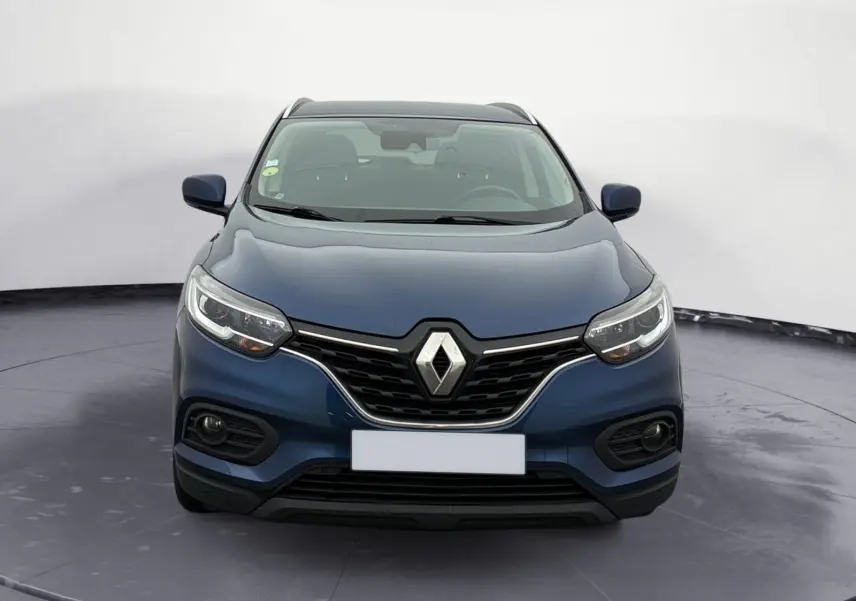 Renault Kadjar bleu cosmos en 3/4 avant droit sur un parking extérieur avec plusieurs fourgons blancs en arrière-plan.