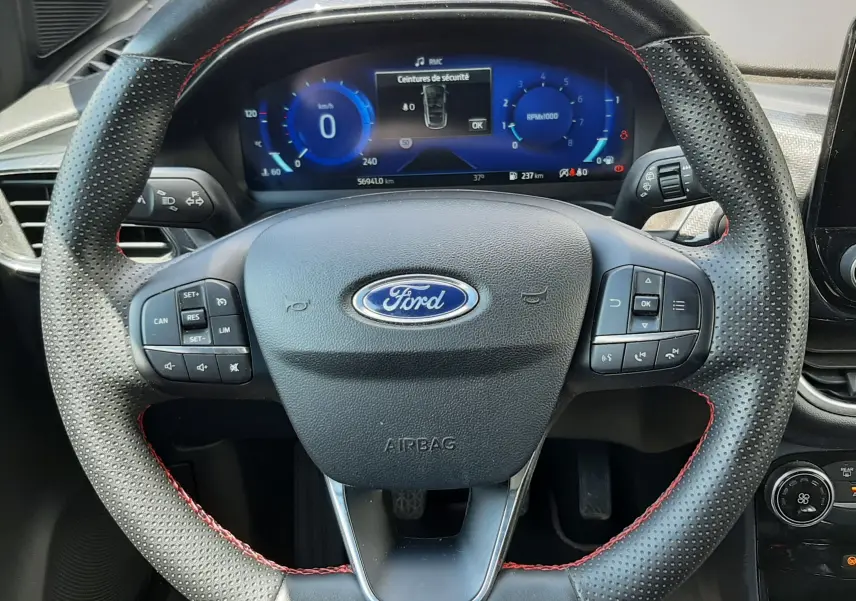Vue rapprochée du volant noir perforé avec surpiqûres rouges du Ford Puma 2022, tableau de bord numérique en arrière-plan.