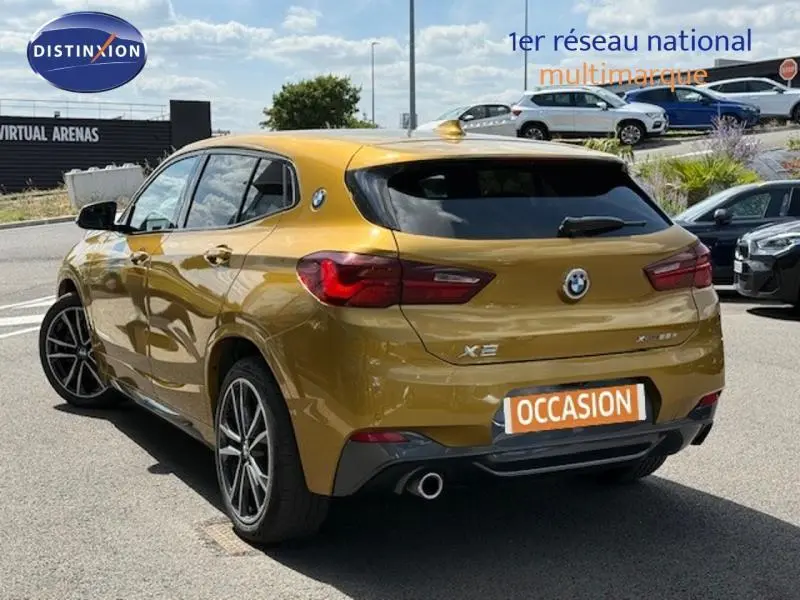 Vue 3/4 arrière droite d'un BMW X2 xDrive25eA 220ch M Sport 2021 en Galvanic Gold métallisé avec logo et échappement visible.
