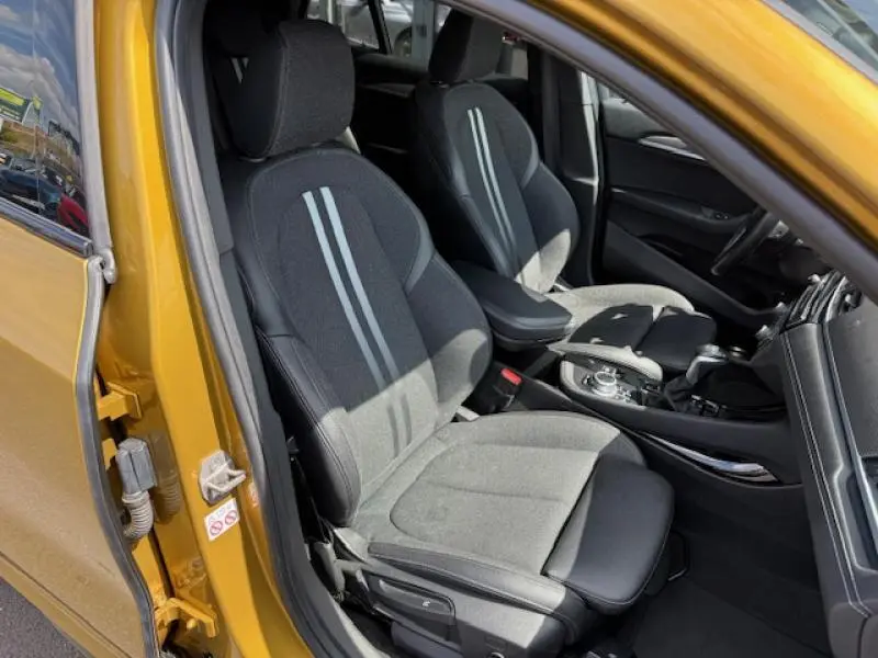 Vue intérieure avant du BMW X2 Galvanic Gold avec sièges sport en tissu gris et détails noirs, porte ouverte côté gauche.