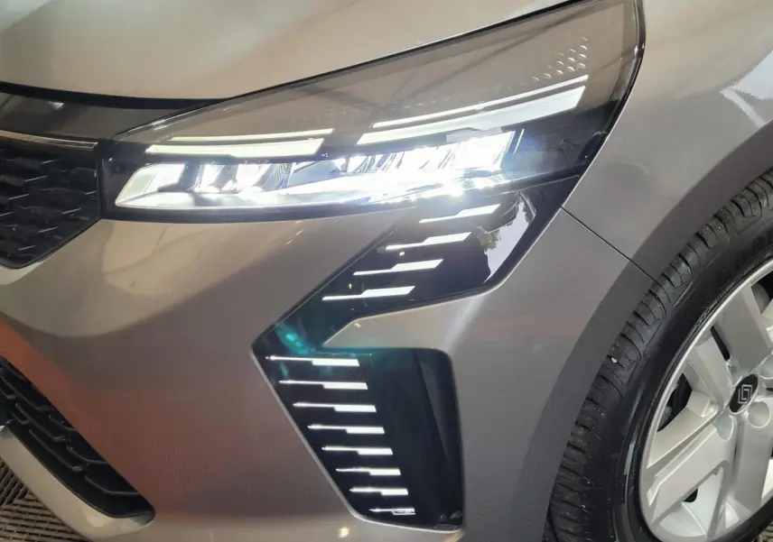 Gros plan sur le phare avant droit allumé d'une Renault Clio gris foncé avec jante alliage et calandre noire.