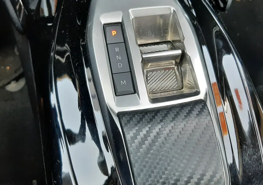 Gros plan sur la console centrale noire brillante du Peugeot 2008 Hybrid 145 Allure avec sélecteur de boîte auto et finition carbone.