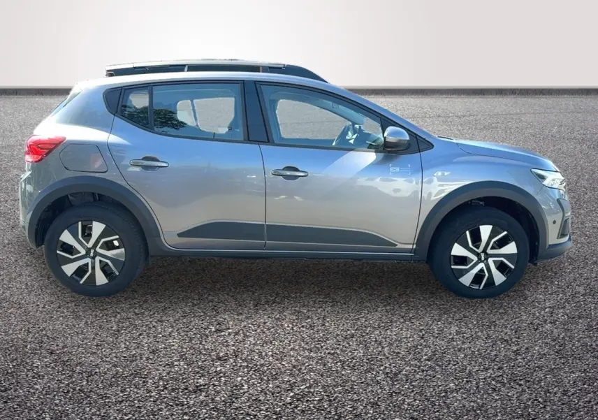 Profil droit d'une Dacia Sandero Stepway gris foncé avec barres de toit et jantes distinctives sur sol gravillonné.