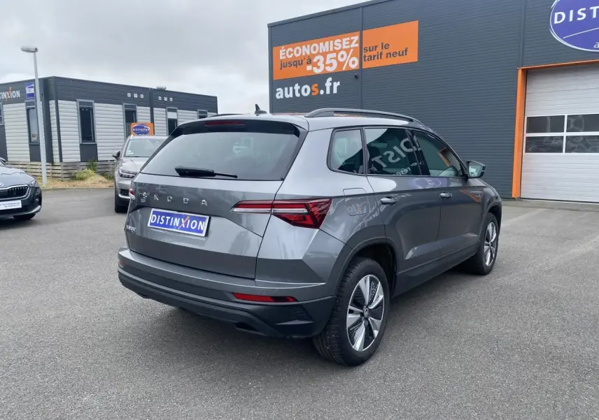 Skoda Karoq gris graphite métallisé vu en 3/4 arrière droit sur parking devant un bâtiment commercial.