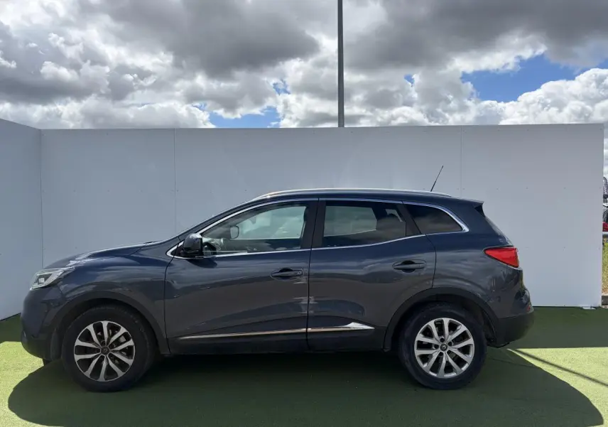 Profil côté gauche d'un Renault Kadjar 2016 gris titanium avec jantes alliage 17 pouces et barres de toit chromées.
