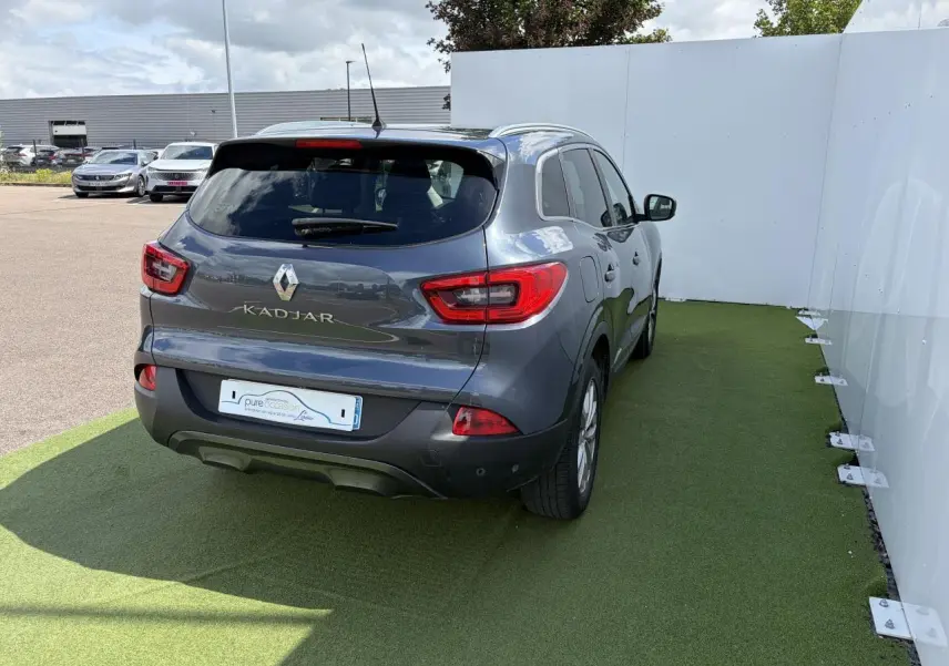 Vue 3/4 arrière droite d'un Renault Kadjar bleu métal garé sur sol vert, avec feux arrière et logo visibles.