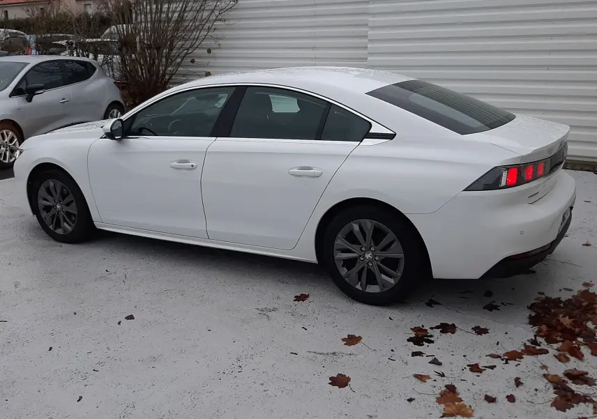 Peugeot 508 blanc vue de profil côté gauche, avec feux arrière allumés et jantes foncées, stationnée sur sol clair.