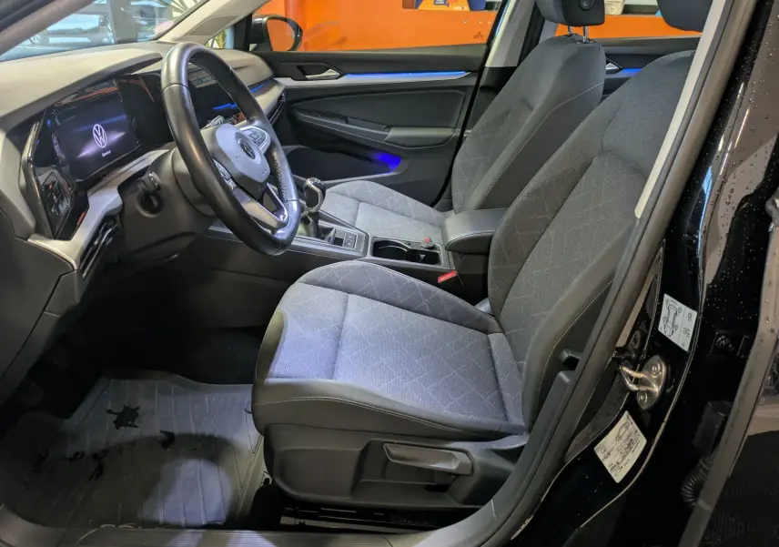 Intérieur avant droit d’une Volkswagen Golf 1.5 eTSI 2024 noire, sièges gris, volant multifonction et éclairage d’ambiance bleu.