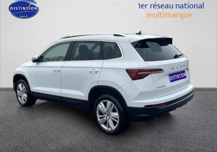 Vue 3/4 arrière droite d'un Skoda Karoq blanc glacial avec jantes alu et barres de toit visibles.