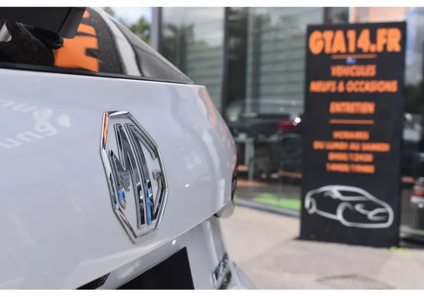 Gros plan sur l'arrière blanc brillant d'une MG3 1.5L Hybrid+ avec logo MG chromé, devant une vitrine commerciale.