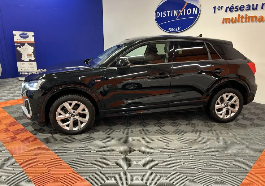 Audi Q2 noir métal vue de profil côté gauche dans un showroom avec jantes argentées et badge S-Line visible.