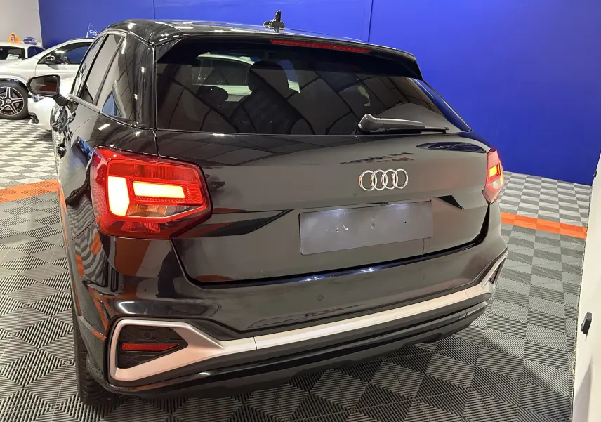 Vue 3/4 arrière d'un Audi Q2 noir métal 2023 avec feux arrière allumés et hayon électrique en intérieur showroom.