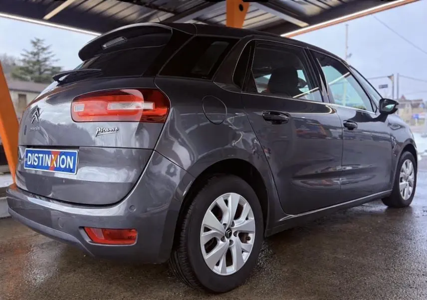 Vue 3/4 arrière droite d'une Citroën C4 Picasso gris platinium métallisé sous abri, avec jantes alliage et feux arrière allongés.