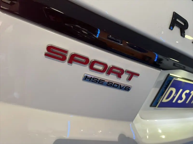 Gros plan sur le badge arrière rouge "SPORT" et "HSE SDV6" du Range Rover Sport blanc, avec partie de la plaque d'immatriculation visible.