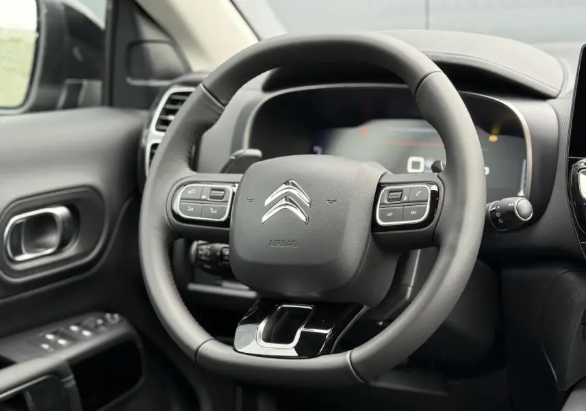 Volant noir de Citroën C5 Aircross Hybrid 2025 vu de face, avec commandes intégrées et tableau de bord numérique flou.