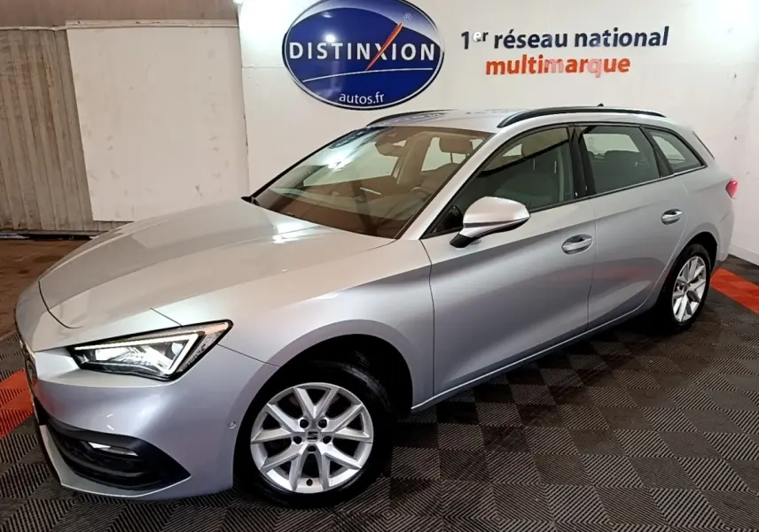 Vue 3/4 avant droit d'une SEAT Leon break gris clair avec jantes alliage et phares LED allumés en intérieur showroom.