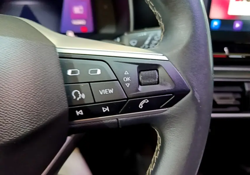 Gros plan sur les commandes à droite du volant cuir noir du SEAT Leon gris foncé, avec écran tactile en arrière-plan flou.
