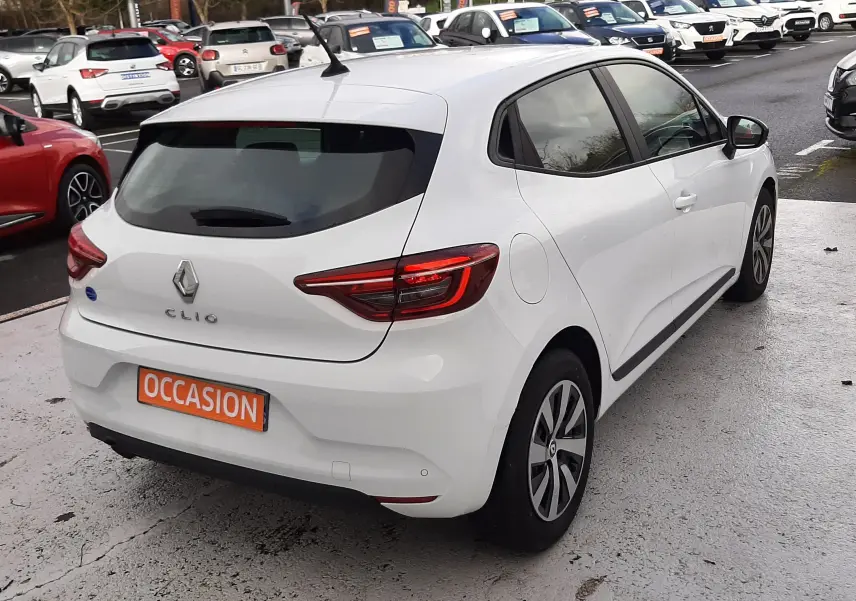 Vue 3/4 arrière droite d'une Renault Clio V blanche 5 portes avec feux LED et plaque "OCCASION" sur parking.
