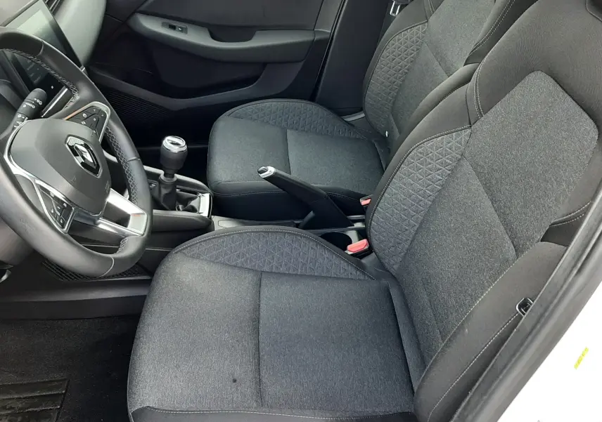 Vue intérieure côté conducteur de la Renault Clio V blanche, sièges tissu noir avec motifs géométriques et boîte manuelle.