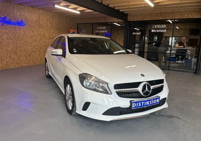 Vue 3/4 avant d'une Mercedes Classe A 200D blanche garée dans un garage avec logo Distinxion sur la plaque avant.