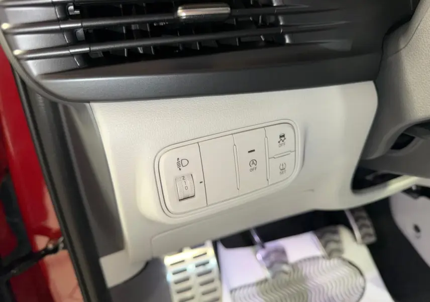 Gros plan sur les commandes de réglage des feux et aides à la conduite, avec pédales sport et intérieur bi-ton gris du Hyundai Bayon rouge.