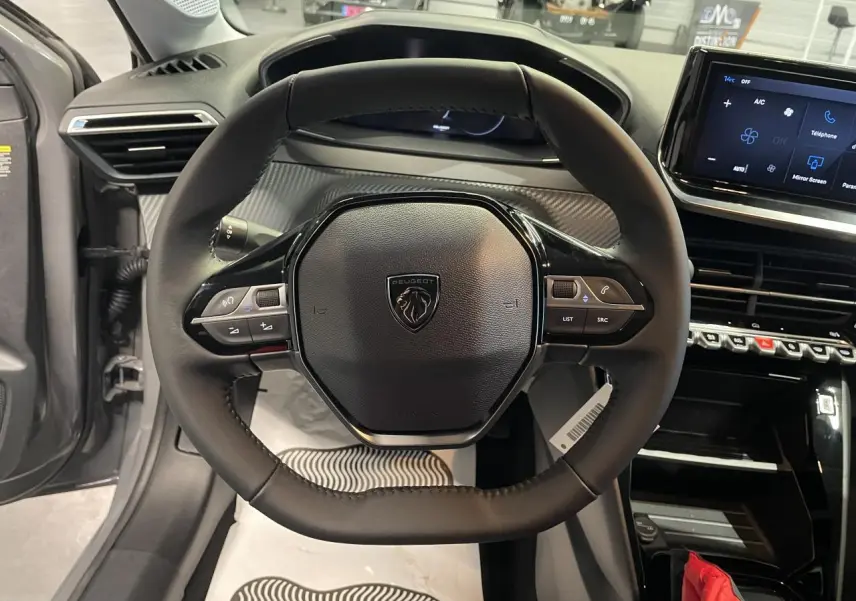 Vue intérieure centrée sur le volant compact cuir de la Peugeot 208 1.2i Hybrid avec tableau de bord numérique.