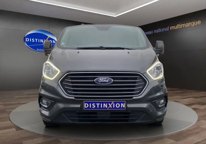 Vue frontale du Ford Tourneo Custom gris Magnetic avec phares allumés et logo Distinxion visible en arrière-plan.