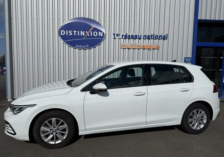Profil droit d'une Volkswagen Golf blanche 1.0 TSI 110 Life de 2020, garée devant un mur avec logo Distinxion.