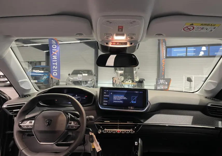 Vue intérieure avant du tableau de bord noir et volant compact cuir de la Peugeot 208 1.2i Hybrid 2025 avec écran tactile central.